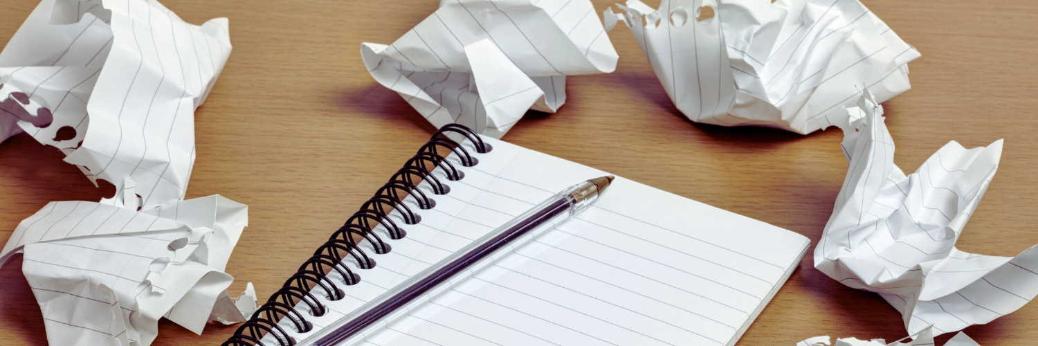 Spiral-bound notebook with blank page, pen placed on top and wadded up pages surrounding it.